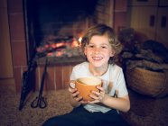 Fröhlicher kleiner Junge sitzt mit Tasse am heimischen Kamin — Stockfoto