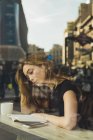 Молода жінка читає книгу в кафе за віконною панеллю з відображенням міста — стокове фото