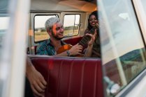 Group of young people laughing and listening to handsome guy playing acoustic guitar inside vintage van in nature — Stock Photo