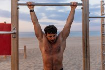 Muscular guy performing pull-ups on bar during sunset on sandy beach — Stock Photo