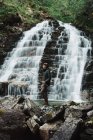 Pescador vestido de chapéu de pesca com camisa azul e calças cinza marchando segurando uma haste giratória em pé sobre rochas atrás da encosta de uma cachoeira — Fotografia de Stock