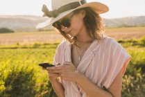 Брюнет в рожевому одязі і елегантний капелюх серфінг смартфон на тлі зеленої літньої сільської місцевості на сонці? — стокове фото