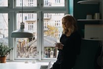 Романтичная блондинка, стоящая с чашкой у окна дома — стоковое фото