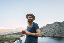 Молодой бородатый мужчина стоит у озера в горах с чашкой и смотрит в камеру — стоковое фото