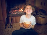 Веселий молодий хлопчик сидить з чашкою в каміні вдома — стокове фото