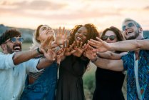 Groupe de jeunes amis divers souriant et tendre les mains vers la caméra tout en se tenant debout sur un fond flou de campagne étonnante pendant le coucher du soleil — Photo de stock