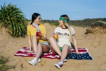 Stylish girlfriends having drinks and sitting on American flag on sand in bright sunshine — Stock Photo