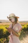 Brunette in pink clothes and elegant hat surfing smartphone on background of green summer countryside in?sunlight — Stock Photo