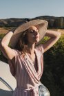 Zarte sinnliche Frau im rosafarbenen Outfit mit elegantem Strohhut und angelehnt an ein Retro-Auto in sommerlicher Landschaft — Stockfoto