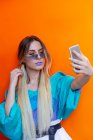 Menina jovem atraente na roupa da moda posando para selfie enquanto está em pé sobre fundo laranja brilhante — Fotografia de Stock