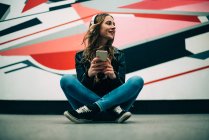 Счастливая молодая женщина сидит на полу с мобильным телефоном и слушает музыку на цветном фоне — стоковое фото
