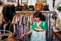 Досить молода жінка посміхається і збирає одяг з рейок одягу, проводячи час у маленькому магазині — стокове фото