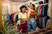 Zwei junge Frauen lächeln und probieren stylische Hüte an, während sie in einem kleinen Geschäft neben einem Spiegel stehen — Stockfoto