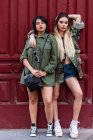 Zwei junge Damen in trendigen Outfits lehnen an einer Hauswand, während sie an einem sonnigen Tag auf der Stadtstraße stehen — Stockfoto