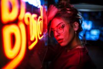 Side view of attractive woman in glasses with eyes closed leaning on wall at lighting neon sign — Stock Photo