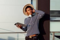 Joven hombre negro con elegante atuendo sosteniendo la tableta y escuchando música mientras se apoya en el pilar del edificio en la calle - foto de stock