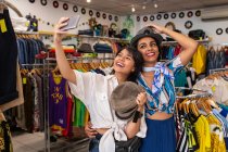 Cheerful friends trying on hat in shop and taking selfie — Stock Photo