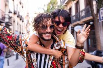 Handsome bearded man in trendy outfit smiling and giving piggyback ride to cheerful girlfriend on blurred background of city street — Stock Photo