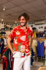 Hombre guapo en traje elegante brillante sonriendo y mirando hacia otro lado mientras está de pie en una pequeña tienda de ropa - foto de stock