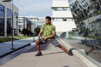Hombre afroamericano calentando piernas en la ciudad con edificios modernos en el fondo - foto de stock