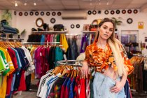 Attraente giovane donna in abito alla moda appoggiata su appendiabiti e guardando la fotocamera mentre in piedi in negozio alla moda — Foto stock