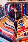 Mulher muito jovem apoiando-se em trilhos de roupas e olhando para a câmera enquanto está de pé em pequena loja — Fotografia de Stock