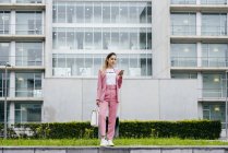 Elegante mujer joven con teléfono inteligente en rosa de pie frente al moderno edificio de oficinas - foto de stock