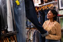 Attractive young lady searching for new outfit on clothes rail in small shop — Stock Photo