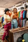 Дві молоді жінки посміхаються і намагаються на стильному капелюсі, стоячи біля дзеркала в маленькому магазині — стокове фото