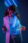 Стильная женщина слушает музыку со смартфоном и наушниками в неоновом свете — стоковое фото