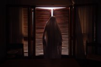 Человек, замаскированный под призрака на Хэллоуин, стоящий в темной комнате — стоковое фото