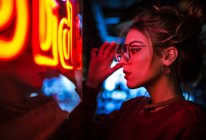 Side view of attractive woman in glasses with eyes closed leaning on wall at lighting neon sign — Stock Photo