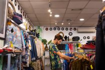 Homem sorrindo e escolhendo vestuário de trilhos de roupas enquanto passa o tempo em pequena loja — Fotografia de Stock