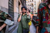 Bella donna asiatica in abito alla moda che tiene gli occhiali da sole e guardando la fotocamera mentre cammina sulla strada della città in mezzo agli amici — Foto stock