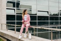 Mulher nova na moda em terno rosa com café e telefone sentado no corrimão em frente ao prédio de escritórios moderno — Fotografia de Stock