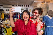 Jovem e mulher em roupas elegantes sorrindo e tirando selfie enquanto está de pé na pequena loja de roupas — Fotografia de Stock