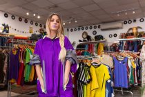 Beautiful young female in bright trendy outfit holding hands in pockets and looking at camera while standing near clothes rail in small store — Stock Photo