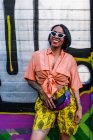 Вид збоку привабливої молодої жінки в модному вбранні сміється і махає рукою, стоячи біля яскравої графіті стіни на вулиці міста — стокове фото