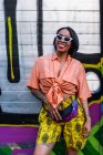 Side view of attractive young female in trendy outfit laughing and waving hand while standing near bright graffiti wall on city street — Stock Photo