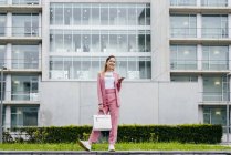 Jeune femme élégante avec smartphone en rose debout devant un immeuble de bureaux moderne — Photo de stock