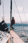 Back view of woman sitting on beak of sailing boat on cold sea water with mountains on background — Foto stock