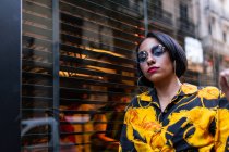 Pretty young female in trendy outfit and sunglasses looking at camera while standing near building window on city street — Stock Photo