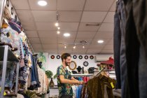 Mann lächelt und pflückt Kleidungsstücke von Kleiderstange, während er Zeit in kleinem Geschäft verbringt — Stockfoto