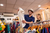 Bonito homem escolhendo camisa elegante enquanto passa o tempo em pequena loja com a namorada — Fotografia de Stock