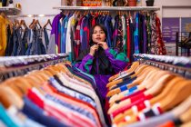 Bella giovane donna appoggiata su binari vestiti e guardando la fotocamera mentre in piedi in un piccolo negozio — Foto stock