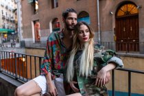 Молодий чоловік і жінка в модному вбранні, сидячи на металевому паркані на вулиці міста і дивлячись на камеру — стокове фото