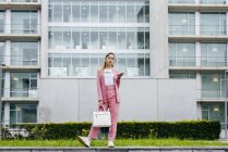 Стильна молода жінка зі смартфоном в рожевому кольорі стоїть перед сучасною офісною будівлею — стокове фото