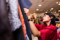 Muito jovem fêmea sorrindo e escolhendo vestuário de trilhos de roupas enquanto passa o tempo em pequena loja — Fotografia de Stock