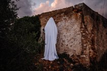 Человек, замаскированный под призрака на Хэллоуин, стоит рядом с разрушенным домом — стоковое фото