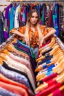 Jolie jeune femme appuyée sur des rails de vêtements et regardant la caméra tout en se tenant dans un petit magasin — Photo de stock